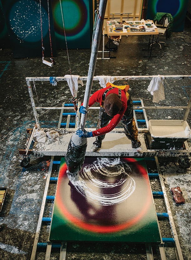 Artist holding a very large paintbrush while standing above the canvas working on an abstract painting in her studio